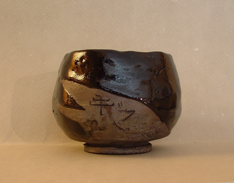 Chawan raku nero modellato a mano, con superfici irregolari e incisione giapponese sulla parete frontale.