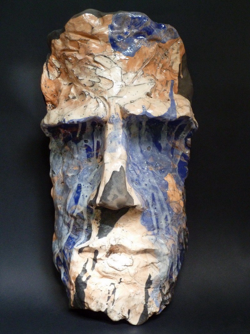 Maschera raku di grandi dimensioni con superfici irregolari, colature cromatiche blu e beige, e un’espressione intensa che suggerisce metamorfosi interiore e memoria ancestrale.
