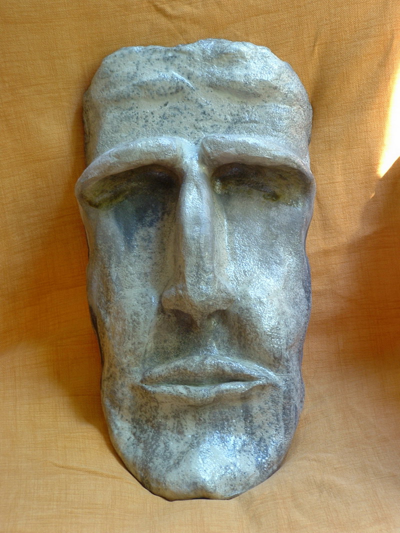 Maschera in argilla raku con superfici fumigate, trama materica irregolare e linee essenziali che evocano un volto primordiale.